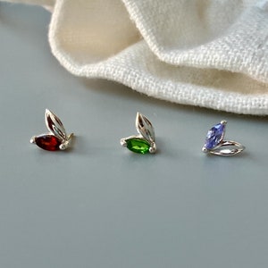 Pode incluir: Três brincos de prata com pedras preciosas de diferentes cores. Os brincos têm a forma de folhas com uma pedra preciosa no centro. As pedras preciosas são vermelha, verde e roxa.