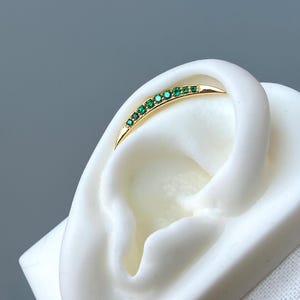 Könnte beinhalten: Ein gebogener, goldfarbener Ohrring mit einer Reihe kleiner, grüner Edelsteine. Der Ohrring ist auf einem weißen, ohrförmigen Display platziert. Die Edelsteine sind gleichmäßig entlang der Kurve des Ohrrings angeordnet.