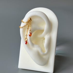May include: A gold-toned ear cuff featuring a small ball stud connected to two delicate chains. Each chain terminates in a small, teardrop-shaped orange crystal charm, creating a layered, dangling effect on the upper ear cartilage.