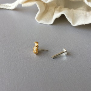 以下が含まれることがあります： 3つの透明な正方形の石が付いた、金色のスタッドピアス。別の銀色のポストも表示されています。ジュエリーは灰色の表面に展示されています。