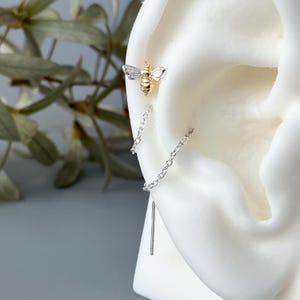 May include: A gold and silver bee-shaped earring with a chain that dangles down. The bee has detailed wings and body. The earring is shown on a white ear model, with a blurred background of green leaves.