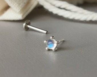 May include: Two silver-colored labret studs. One stud has a flat disc backing and a straight post. The other stud features a small, round, iridescent gemstone set in a star-shaped bezel. The gemstone has blue and white hues.