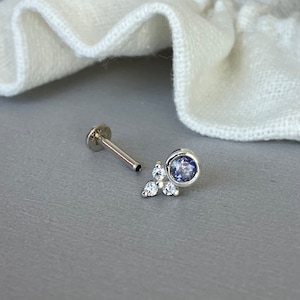 Peut inclure: Un bijou de lèvre en argent avec un petit dos plat et rond et une tige fine. Il présente une pierre précieuse bleu clair sertie dans un sertissage et trois petites pierres précieuses claires en grappe.
