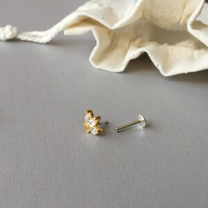 以下が含まれることがあります： クリアな輝きの石が特徴の、花のモチーフが付いたゴールドカラーのスタッドピアス。 ピアスはシルバーのポストと小さなオフホワイトの巾着袋の隣にあり、すべてグレーの背景に配置されています。
