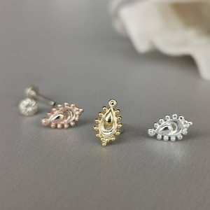 May include: Three ornate stud earrings in rose gold, gold, and silver. Each earring features a teardrop design with intricate detailing. The earrings are displayed on a gray surface, showcasing their delicate craftsmanship.