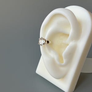 May include: A silver ear cuff with a clear, faceted gemstone. The cuff is worn on a white mannequin ear.