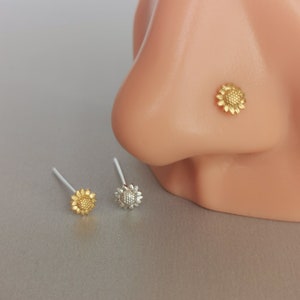 Puede incluir: Tres anillos de nariz de plata con un diseño de girasol. El girasol es de color dorado. Los anillos de nariz están sobre una superficie blanca.