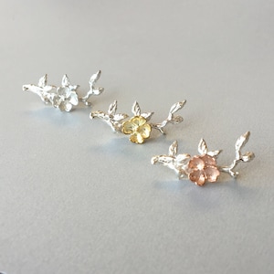May include: Three floral branch-shaped jewellery pieces. One is silver, one has a gold flower, and the third has a rose gold flower. Each piece has a flower and several leaves. The jewellery is displayed on a light grey surface.