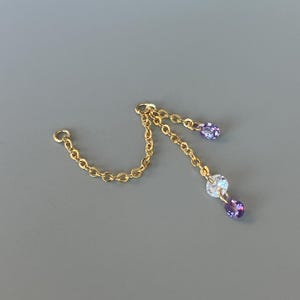 May include: A gold-coloured chain with a small loop at one end and a decorative chain with two purple teardrop-shaped gems and one clear gem. The jewellery is set against a grey background.
