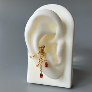 May include: A gold-colored ear piercing with dangling chains and three teardrop-shaped, orange-red gemstones. The earring is displayed on a white ear model against a gray background. The jewelry is designed to be worn on the helix.