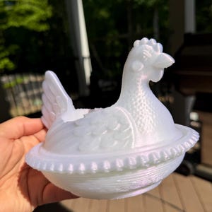 May include: A white hen-shaped dish with a lid, likely for decorative or kitchen use. The hen has detailed feather and comb textures. The dish is held in a hand, with a blurred outdoor background.