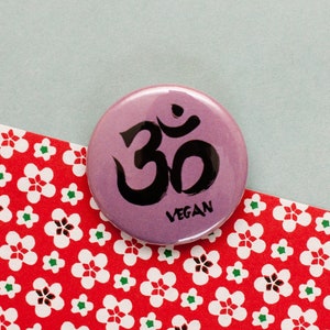 Puede incluir: Un botón rosa con un símbolo Om negro y la palabra "Vegan" escrita debajo. El botón está sobre un fondo floral rojo y blanco.