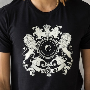 Puede incluir: Una camiseta negra con un gráfico dorado de un escudo de armas con dos leones, un altavoz y la palabra "JUNGLIST".