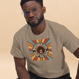 Puede incluir: Una camiseta de color marrón claro con un gráfico retro de una mujer con afro que lleva gafas de sol. El gráfico dice "Northern Soul Keep The Faith" en un círculo con rayos de colores que irradian hacia afuera.