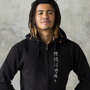 May include: Black zip-up hoodie with white Japanese text on the front. The hoodie has a drawstring hood and a kangaroo pocket. The model is wearing the hoodie. The text reads "継続は力なり".