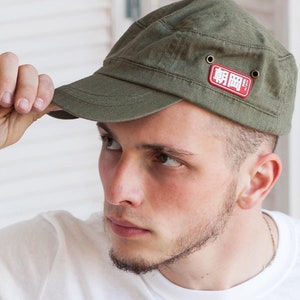 May include: A man wearing a white t-shirt with gold kanji characters printed on the front. He is wearing a green military style cap with a red patch that says "戦闘" in white lettering.