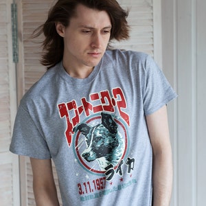 Könnte beinhalten: Ein graues T-Shirt mit einem Grafikdruck eines Hundekopfes in einem Kreis mit dem Text "ライカ" und "3.11.1957" in Japanisch. Der Text "世界初! 宇宙船に乗った犬" ist ebenfalls auf dem Shirt gedruckt.
