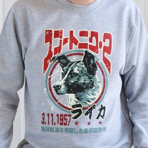Puede incluir: Una sudadera gris con un estampado gráfico de un perro en un círculo con el texto "Спутник-2" y "3.1.1957 Лайка" en caracteres japoneses. El texto debajo del círculo dice "地球軌道を周回した最初の生物".