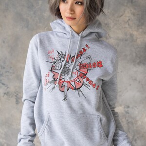 Puede incluir: Una sudadera gris con capucha con un estampado gr&aacute;fico de un personaje de anime dando una patada con el texto "Fight!" y texto japon&eacute;s. El personaje lleva una falda y tiene el pelo largo.