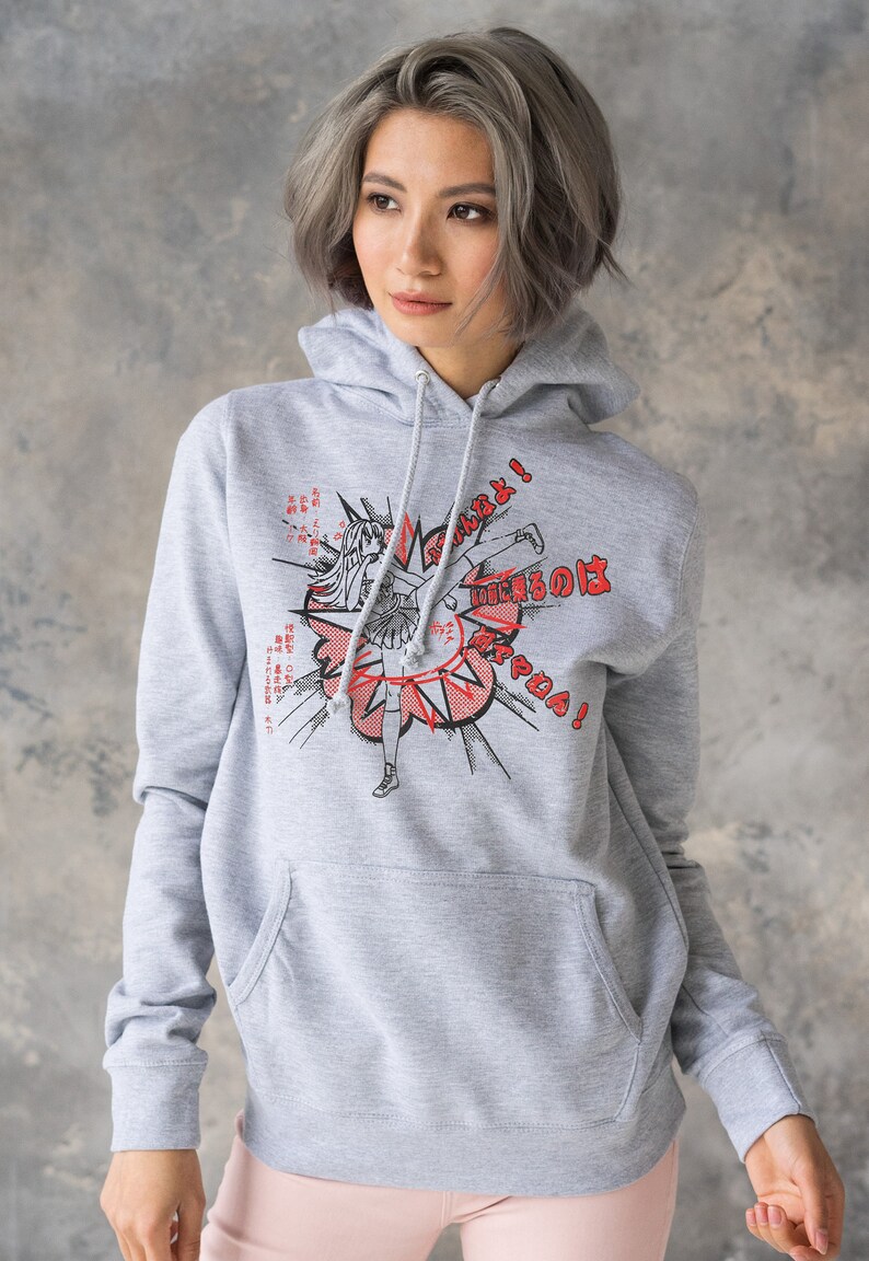 Puede incluir: Una sudadera gris con capucha con un estampado gr&aacute;fico de un personaje de anime dando una patada con el texto "Fight!" y texto japon&eacute;s. El personaje lleva una falda y tiene el pelo largo.