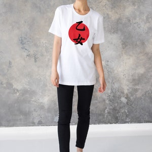 May include: A white t-shirt with a red circle and black Japanese characters that say "乙 女" on the front. The person wearing the shirt is standing in front of a gray concrete wall.