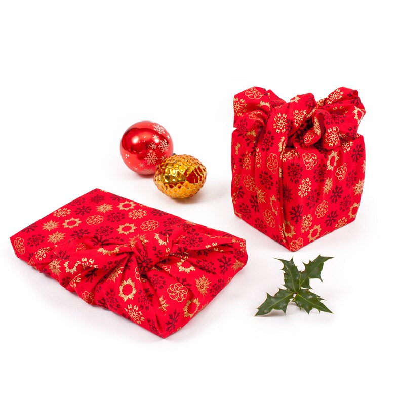 May include: Two red and gold gift boxes wrapped in fabric with a snowflake pattern. The boxes are tied with fabric and are sitting on a white surface. There are three Christmas ornaments, two red and one gold, in the background.