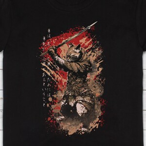 May include: A black t-shirt with a graphic of a cat wielding a sword in a warrior pose. The cat is surrounded by red and brown splatters of paint. The Japanese text on the shirt reads "The warrior's heart is like a cat's, it is always ready to fight."