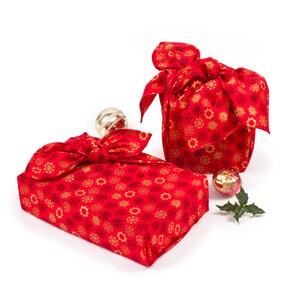 May include: Two red gift boxes wrapped in fabric with a snowflake pattern. The boxes are tied with a red ribbon and have gold accents. The boxes are sitting on a white surface.