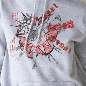Puede incluir: Sudadera con capucha gris con un estampado gr&aacute;fico de una chica de dibujos animados dando una patada con el texto "はなやねん!" y texto japon&eacute;s en rojo y negro.