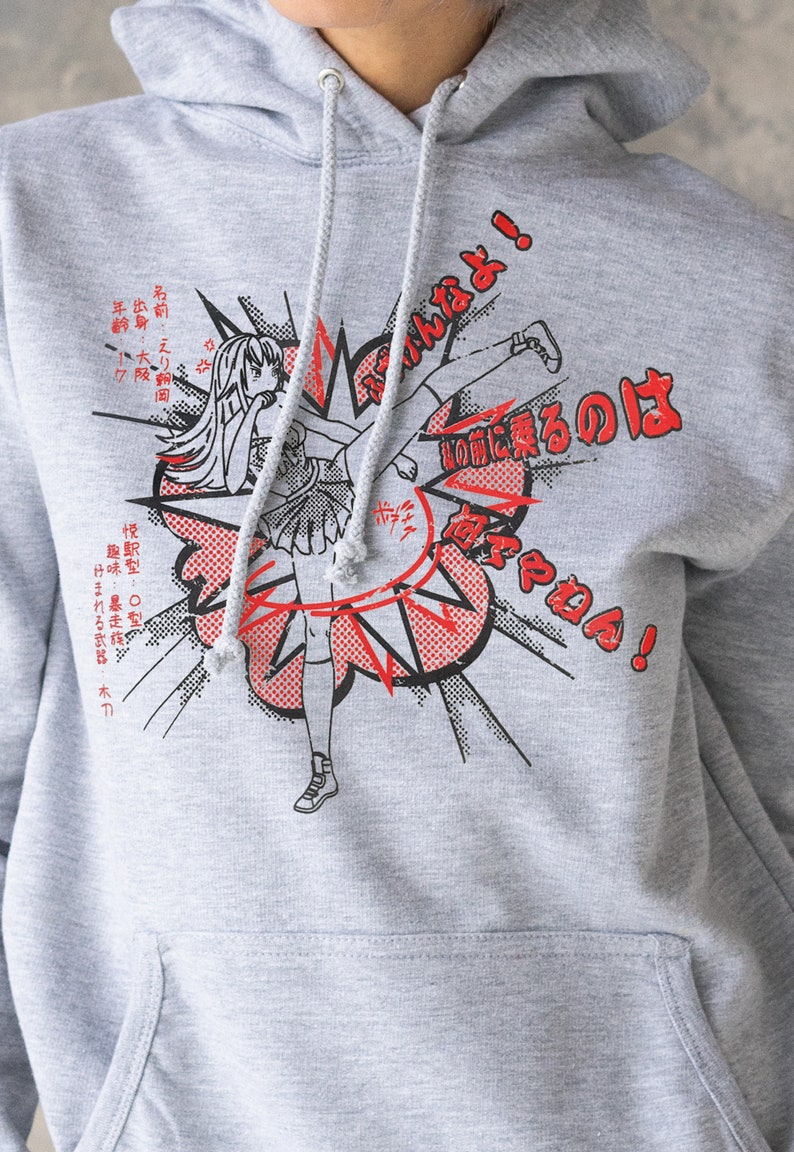 Puede incluir: Sudadera con capucha gris con un estampado gr&aacute;fico de una chica de dibujos animados dando una patada con el texto "はなやねん!" y texto japon&eacute;s en rojo y negro.
