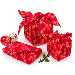 May include: Three red gift boxes wrapped in fabric with a gold snowflake pattern. The boxes are of different sizes and are arranged on a white surface. There are two red and white Christmas ornaments in the background.