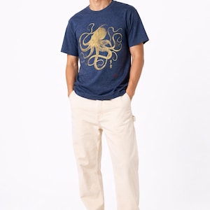 Pode incluir: Uma pessoa vestindo uma camiseta azul marinho com um desenho de polvo dourado e cal&ccedil;as cor creme. A camiseta tem gola redonda e mangas curtas. A pessoa tamb&eacute;m est&aacute; usando t&ecirc;nis preto e branco.