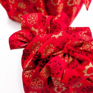 May include: Red fabric gift wrap with a gold snowflake pattern. The fabric is tied in a knot and the ends are hanging loose.