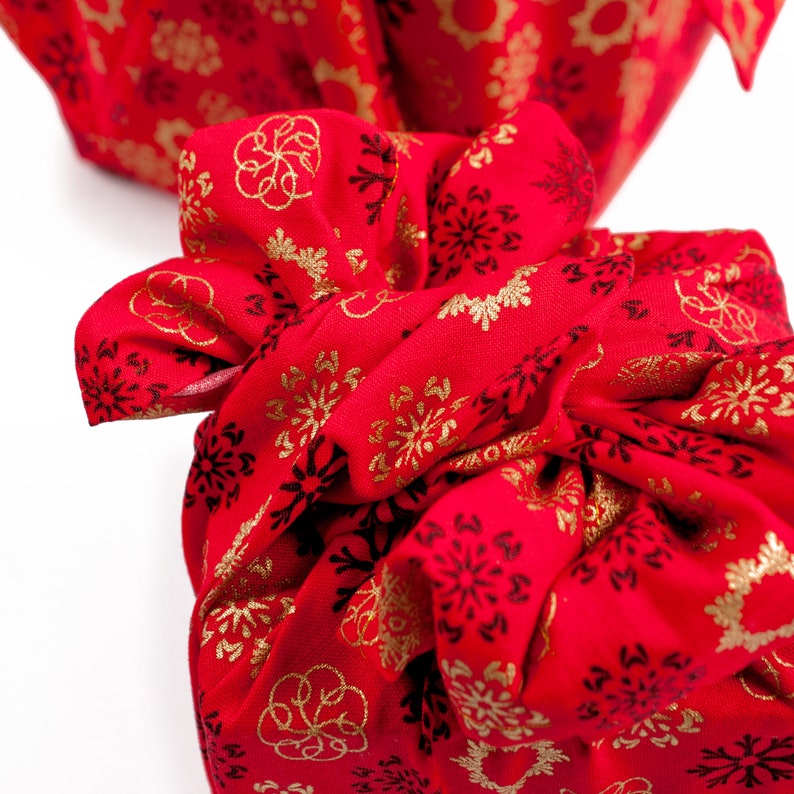 May include: Red fabric gift wrap with a gold snowflake pattern. The fabric is tied in a knot and the ends are hanging loose.