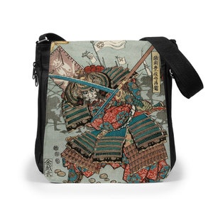 May include: A black crossbody bag featuring a printed panel of a Japanese samurai in traditional armor holding a sword. The artwork includes red, blue, and green patterns, with Japanese text characters visible on the right and bottom left sides.