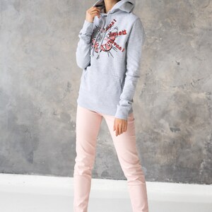 Puede incluir: Una sudadera gris con capucha con un estampado gr&aacute;fico de una mujer con un vestido rojo y el texto "Superwoman!" en rojo. La modelo lleva la sudadera con pantalones rosas y zapatillas blancas.