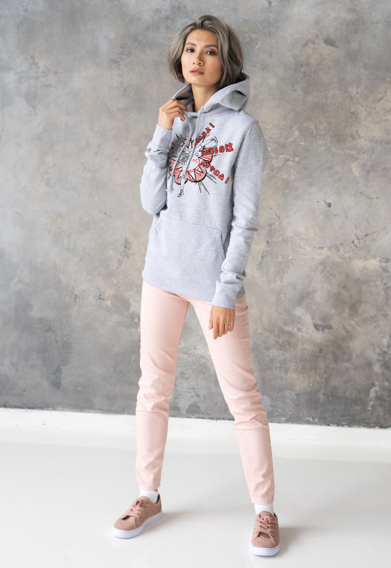 Puede incluir: Una sudadera gris con capucha con un estampado gr&aacute;fico de una mujer con un vestido rojo y el texto "Superwoman!" en rojo. La modelo lleva la sudadera con pantalones rosas y zapatillas blancas.