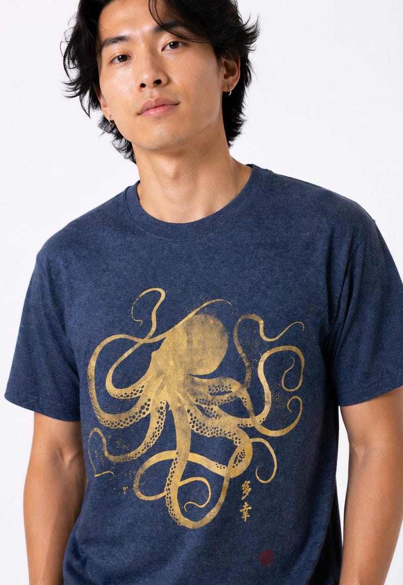 Pode incluir: Camiseta azul marinho com um desenho de polvo dourado. A ilustra&ccedil;&atilde;o do polvo apresenta tent&aacute;culos em espiral e um corpo texturizado. A camiseta tem gola redonda e mangas curtas. Caracteres japoneses s&atilde;o vis&iacute;veis.