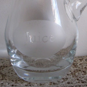 Peut inclure: Un pichet en verre transparent avec un ovale givré sur le devant qui dit "Jus". Le pichet a une poignée et une base arrondie.