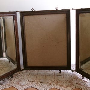 May include: A vintage, three-panel vanity mirror with a dark brown wooden frame. The mirror has a central panel for a photo and two side panels with mirrors. The mirror is set on small, decorative feet.