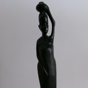 Könnte beinhalten: Eine schwarze Holzskulptur einer Frau, die einen Topf auf dem Kopf trägt. Die Skulptur ist in einfacher, stilisierter Weise geschnitzt und hat ein traditionelles afrikanisches Flair.