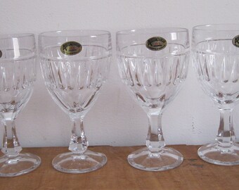 May include: Four clear crystal wine glasses with a decorative etched pattern. Each glass has a stem and a round base. The glasses are arranged in a row on a wooden surface.
