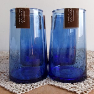May include: Two cobalt blue Moroccan glassware tumblers with paper tags. The cylindrical glasses have a slightly flared base and are displayed on a cream-colored, crocheted doily. The tags read "MOROCCAN GLASSWARE, From Culture."