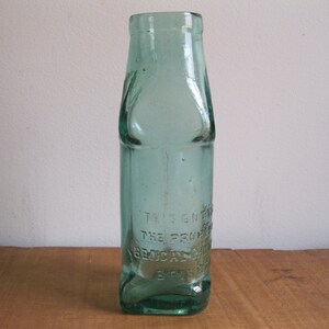 Peut inclure: Une bouteille en verre vert clair avec une surface texturée. La bouteille a un col étroit et une base large. Le texte "THIS BOTTLE THE PROPERTY OF BEVERAGE CO. ST. LOUIS, MO." est gravé sur la bouteille.