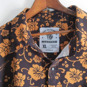 May include: Brown button-up shirt with a floral pattern in orange. The shirt is hanging on a wooden hanger. The shirt has a label that says "SAVTAC" and "AGE TACTICIAN". The shirt is size XL.