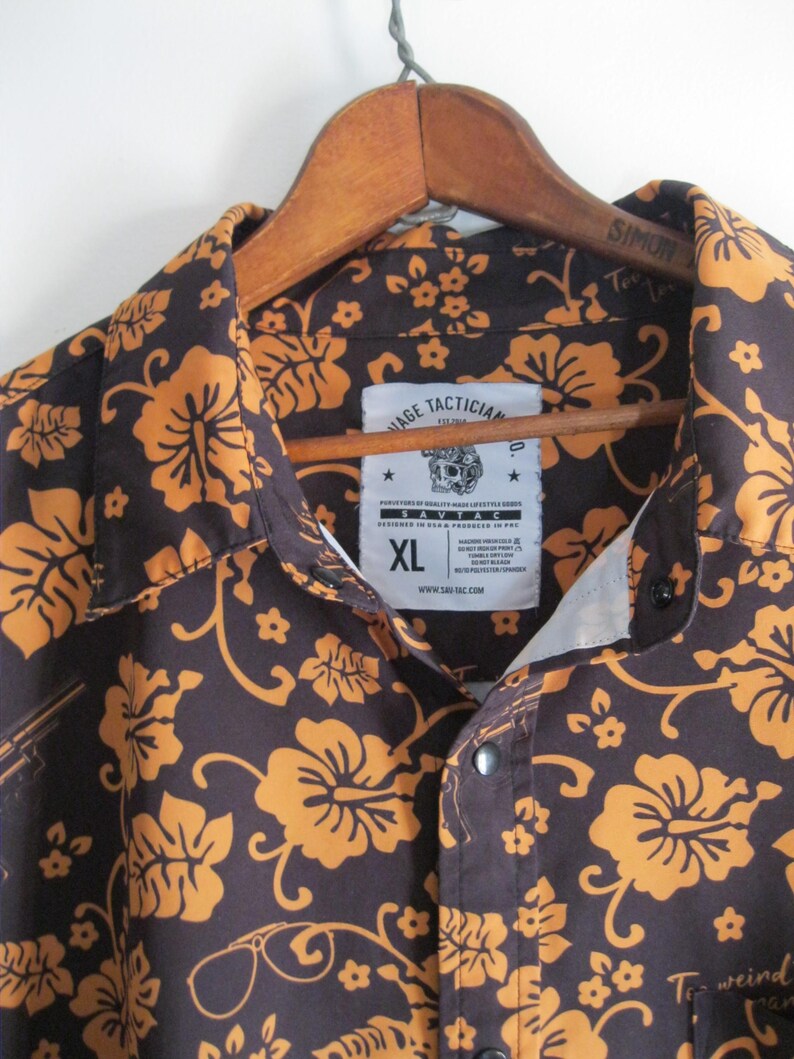 May include: Brown button-up shirt with a floral pattern in orange. The shirt is hanging on a wooden hanger. The shirt has a label that says "SAVTAC" and "AGE TACTICIAN". The shirt is size XL.