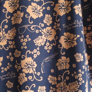 May include: Navy blue fabric with a repeating pattern of tan hibiscus flowers, leaves, and small white flowers. The design includes a revolver, eyeglasses, and the text "Too weird to live, too rare to die."