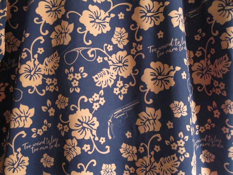 May include: Navy blue fabric with a repeating pattern of tan hibiscus flowers, leaves, and small white flowers. The design includes a revolver, eyeglasses, and the text "Too weird to live, too rare to die."
