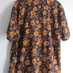 May include: Dark brown short-sleeve shirt with a floral pattern in shades of orange. The shirt features a button-down collar and a relaxed fit. The design includes sunglasses and text that reads "Too proud to live, too mean to die."