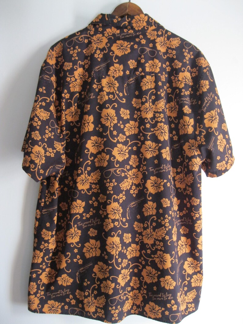 May include: Dark brown short-sleeve shirt with a floral pattern in shades of orange. The shirt features a button-down collar and a relaxed fit. The design includes sunglasses and text that reads "Too proud to live, too mean to die."